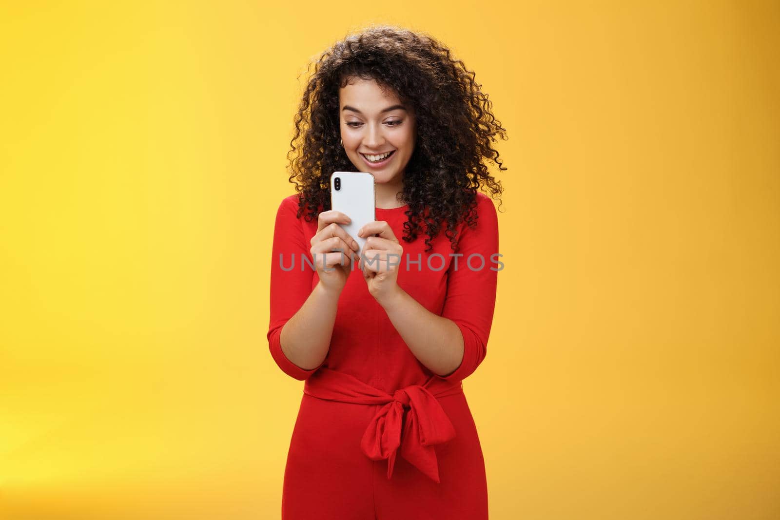 Wow new mobile phone amazing. Impressed and astonished good-looking curly-haired female in red dress holding smartphone looking at screen amused as playing cool app or game over yellow wall by Benzoix