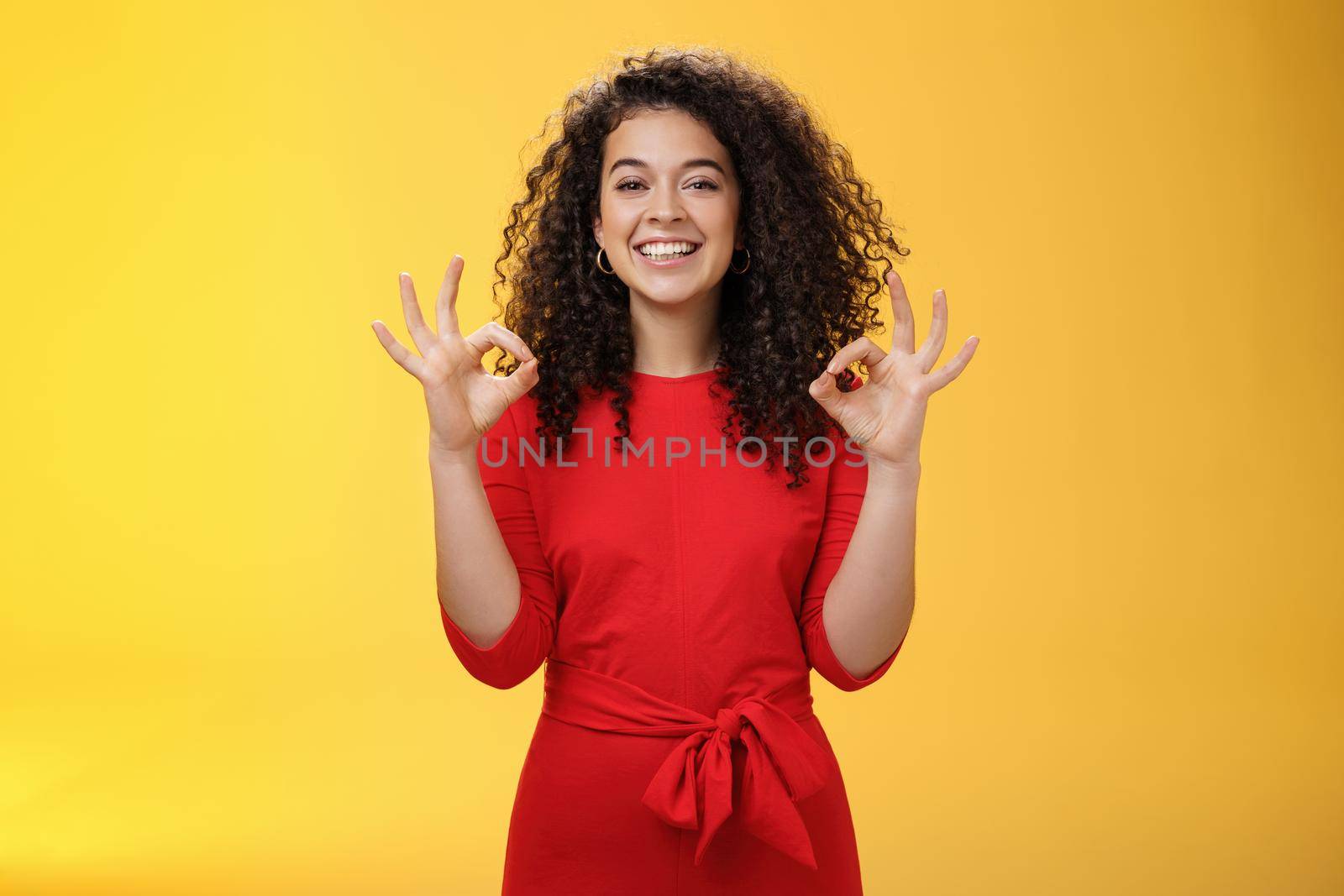 Girl pleased everything goes on plan and okay. Happy joyful charismatic woman with curly hair in red dress showing ok gesture in approval and like, satisfied with good work over yellow wall by Benzoix