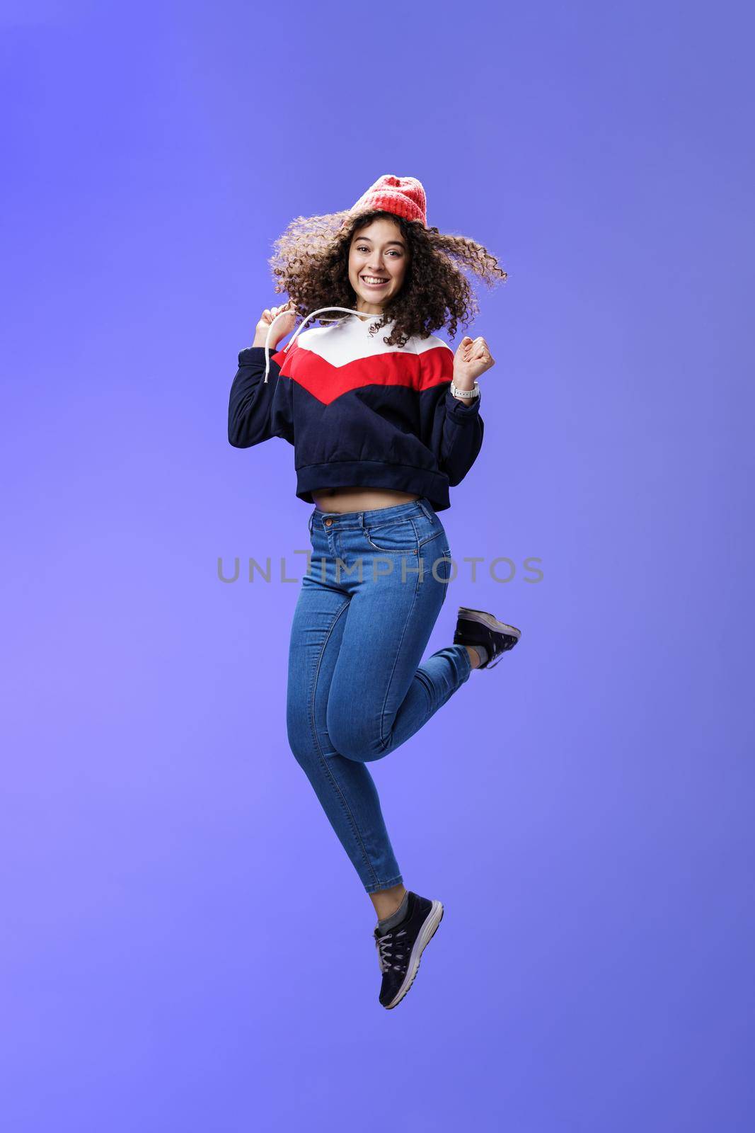 Delighted carefree and happy young woman with curly hair in warm beanie jumping joyfully having fun raising hands and smiling broadly posing over blue background in outdoor clothes.