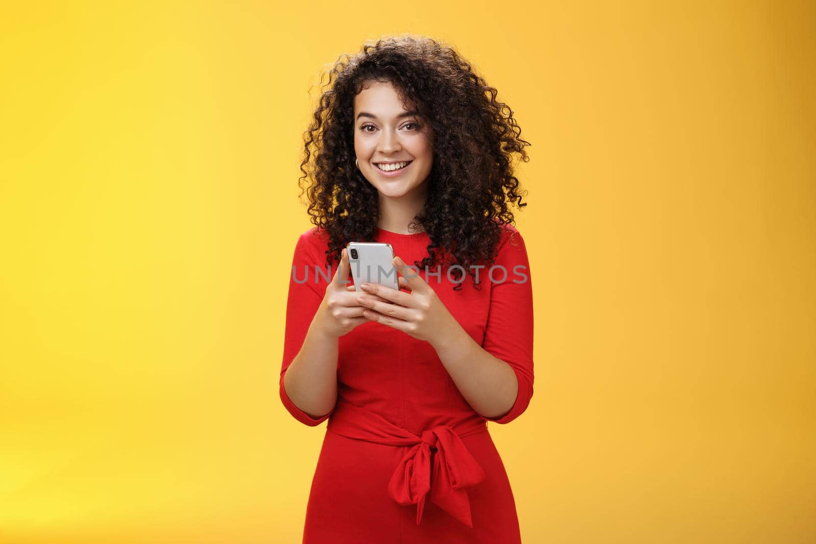 Girl messaging friend to tell all hot rumors holding mobile phone in hands smiling broadly and excited at camera as using smartphone browsing in net or edditing in app over yellow background. Technology concept