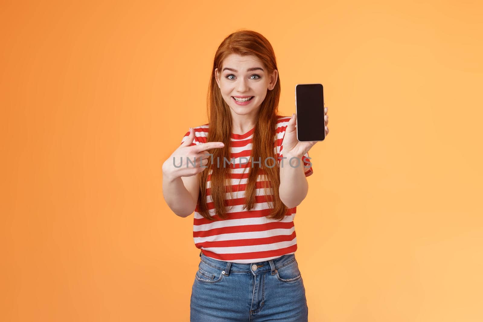 Impressed excited happy redhead woman smiling broadly, hold smartphone, pointing cellphone screen, thrilled amazed introduce fascinating application, stand orange background rejoice.
