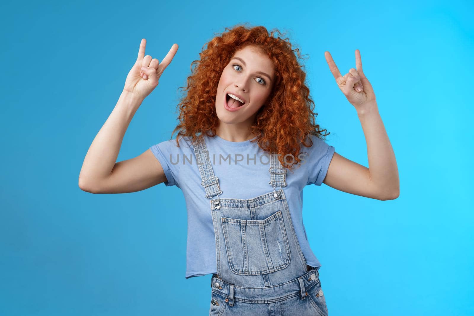 Daring cool stylish awesome redhead cheerful curly-haired girl tilt head show tongue joyfully stare camera playful raise hands rock-n-roll heavy metal gesture having fun blue background.