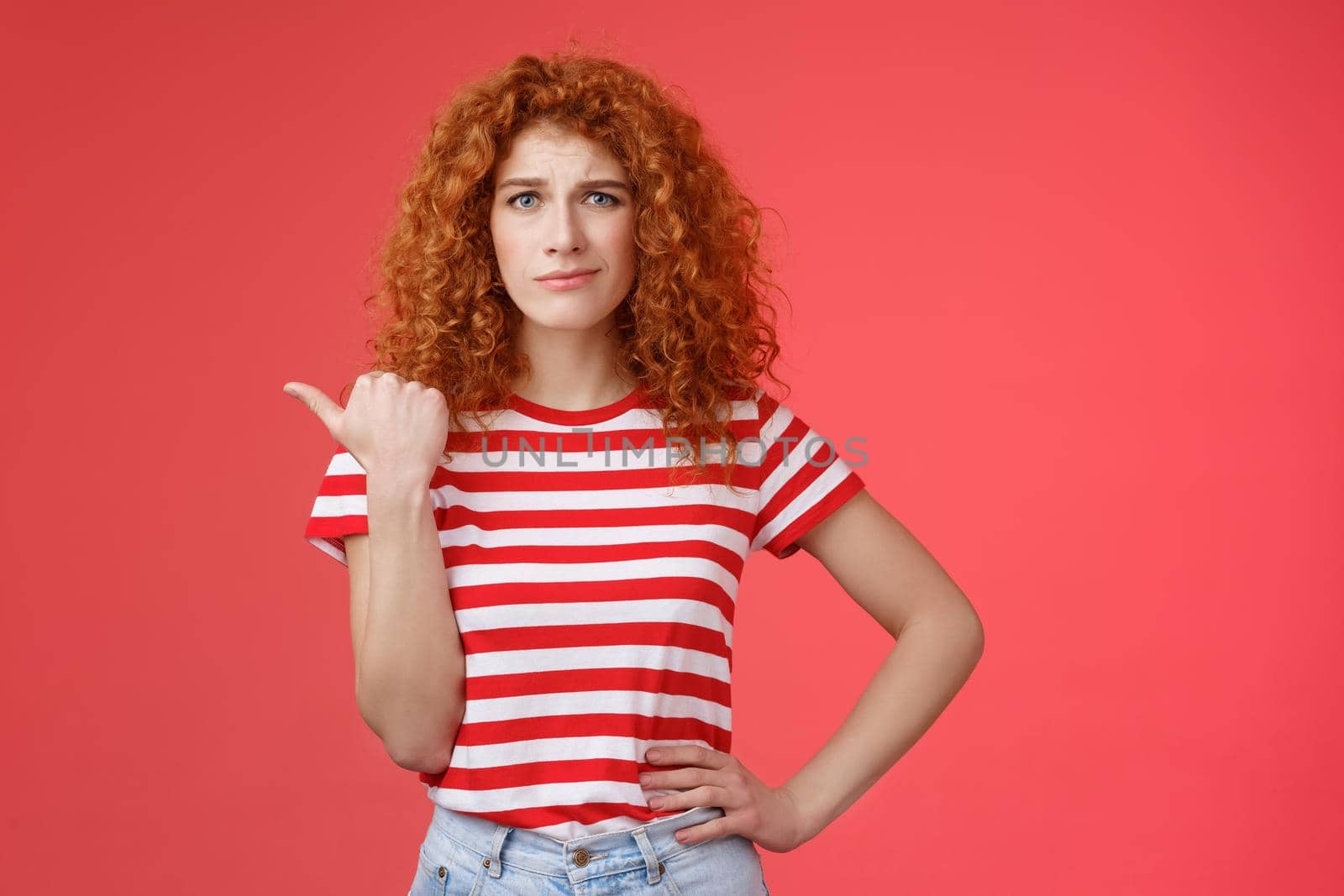 Girl not sure best choice. Doubtful redhead curly woman look suspicious frowning uncertain stare hesitant pointing left thumb unsure buy or not standing red background displeased by Benzoix