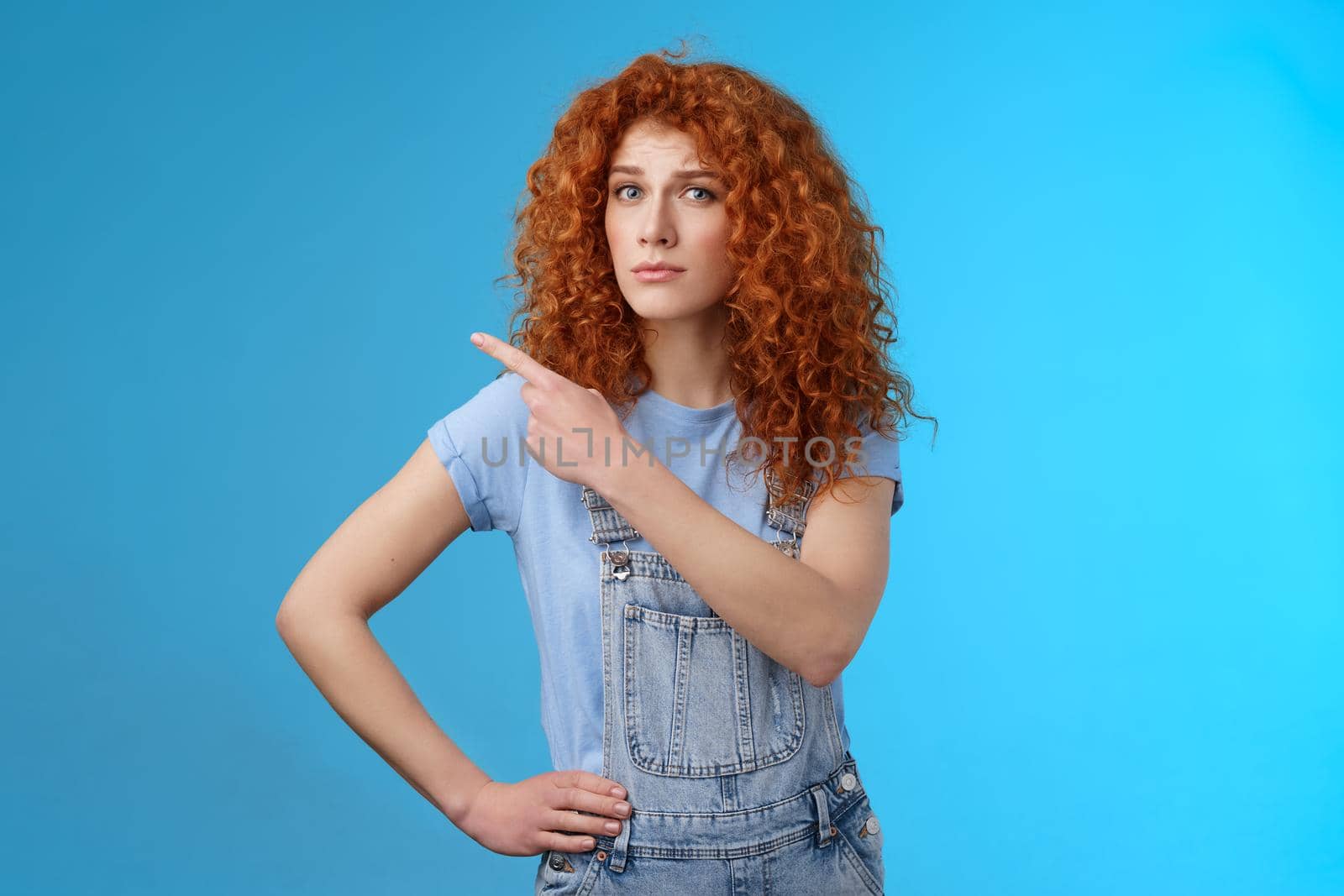 Unsure silly timid hesitant cute redhead curly-haired ginger girl frowning uncertain asking your opinion questioned look camera pointing upper left corner confused worried blue background.