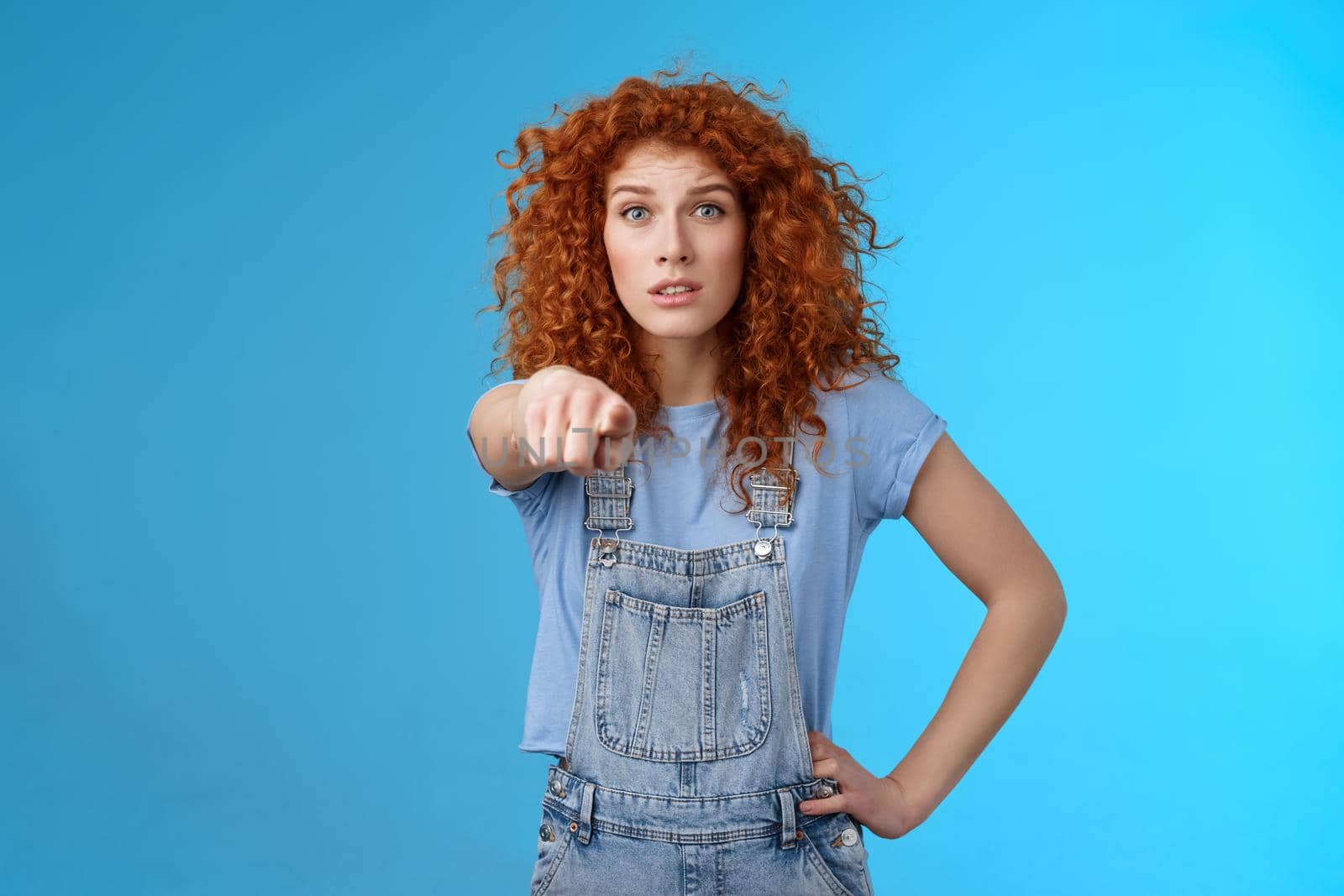 You seem familiar. Worried concerned lively redhead emotive curly girl pointing camera look excited nervously recognize someone standing confused hold hand waist wear overalls. Copy space