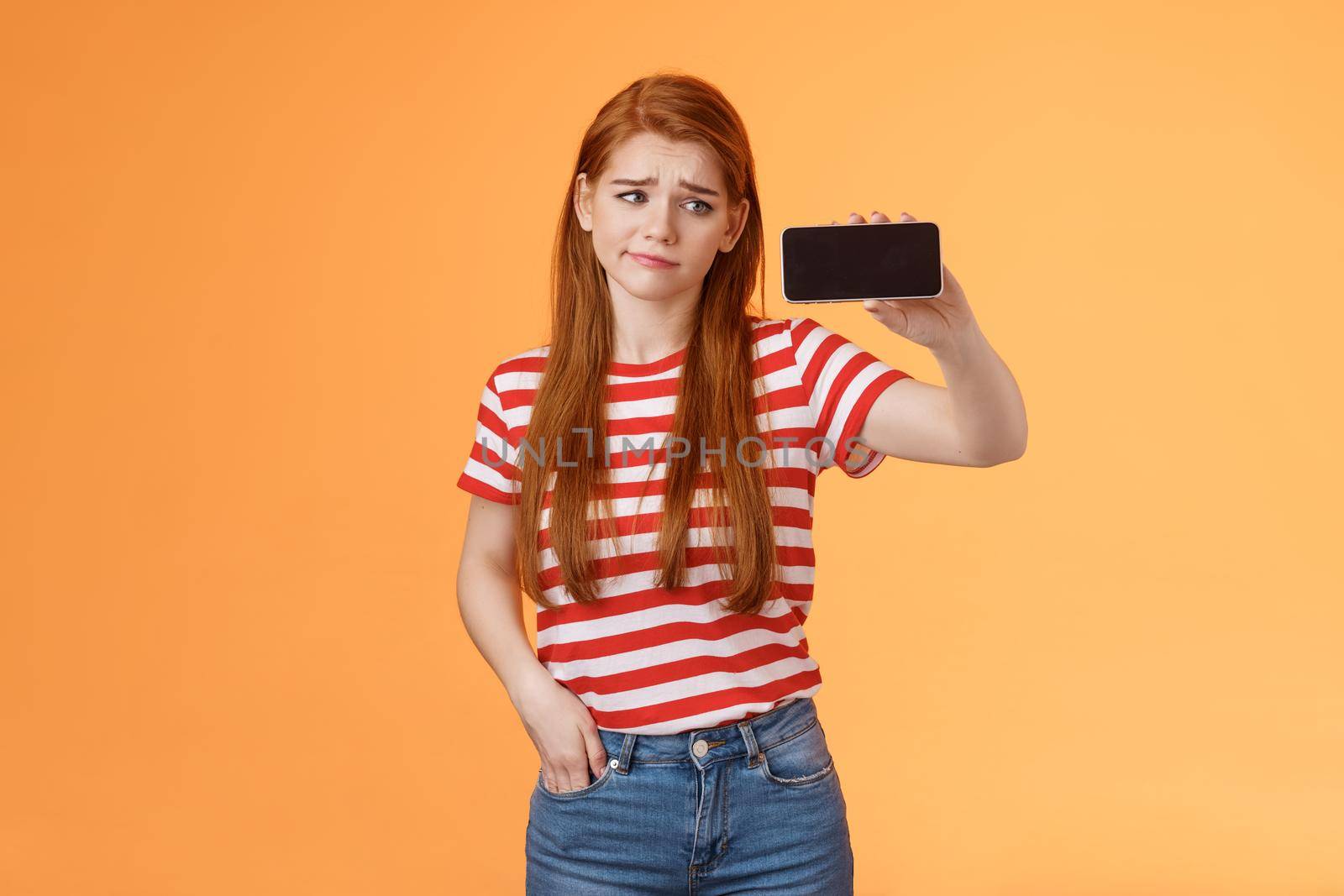 Hesitant disappointed cute redhead woman smirk displeased, hold smartphone horizontal, look device upset, doubtful about blog popularity, sigh uneasy, stare gadget perplexed, show telephone screen.