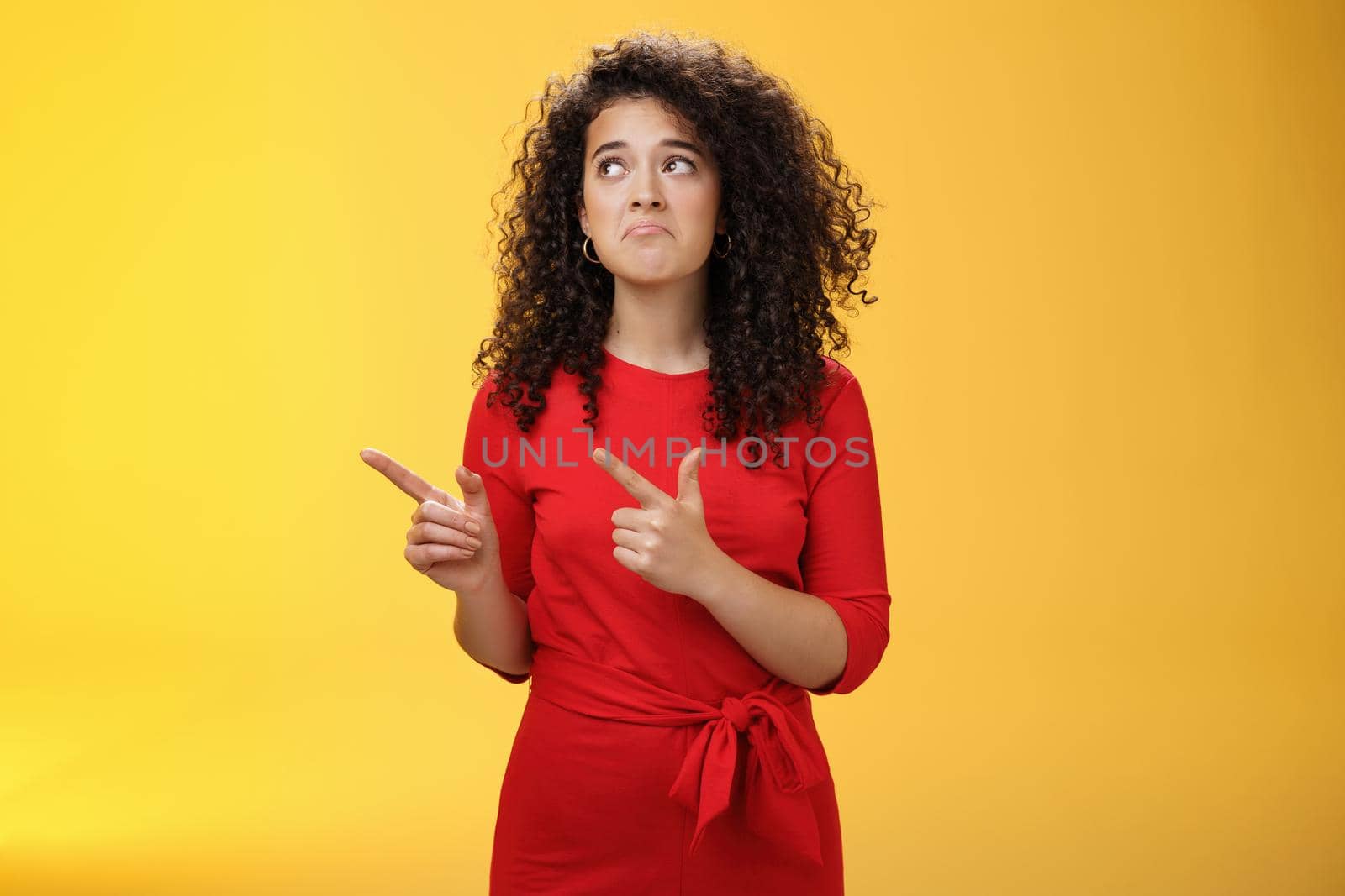 Gloomy upset and sad cute silly curly-haired girl in red dress pursing lips down in unhappy smile frowning from regret, missing chance or offer pointing at upper left corner over yellow background.