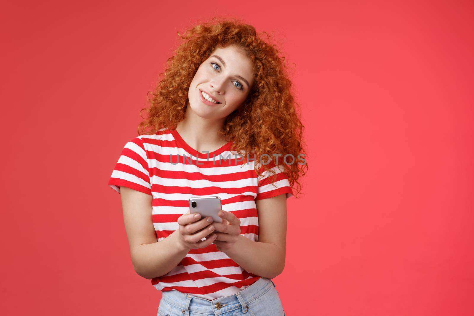 Silly cute redhead curly-haired fashionable female hold smartphone tilt head smiling broadly toothy positive grin pondering what answer send provocative sassy message red background.