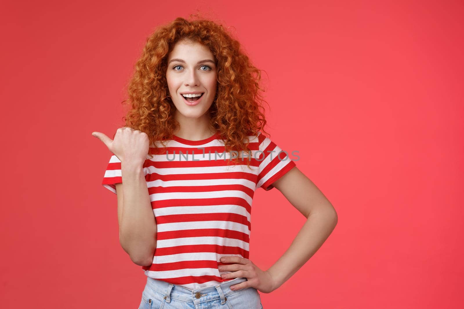 Sassy good-looking emotive happy smiling redhead european female curly hairstyle pointing thumb left grin assertive cheeky hold hand waist directing promo advertising offer red background.