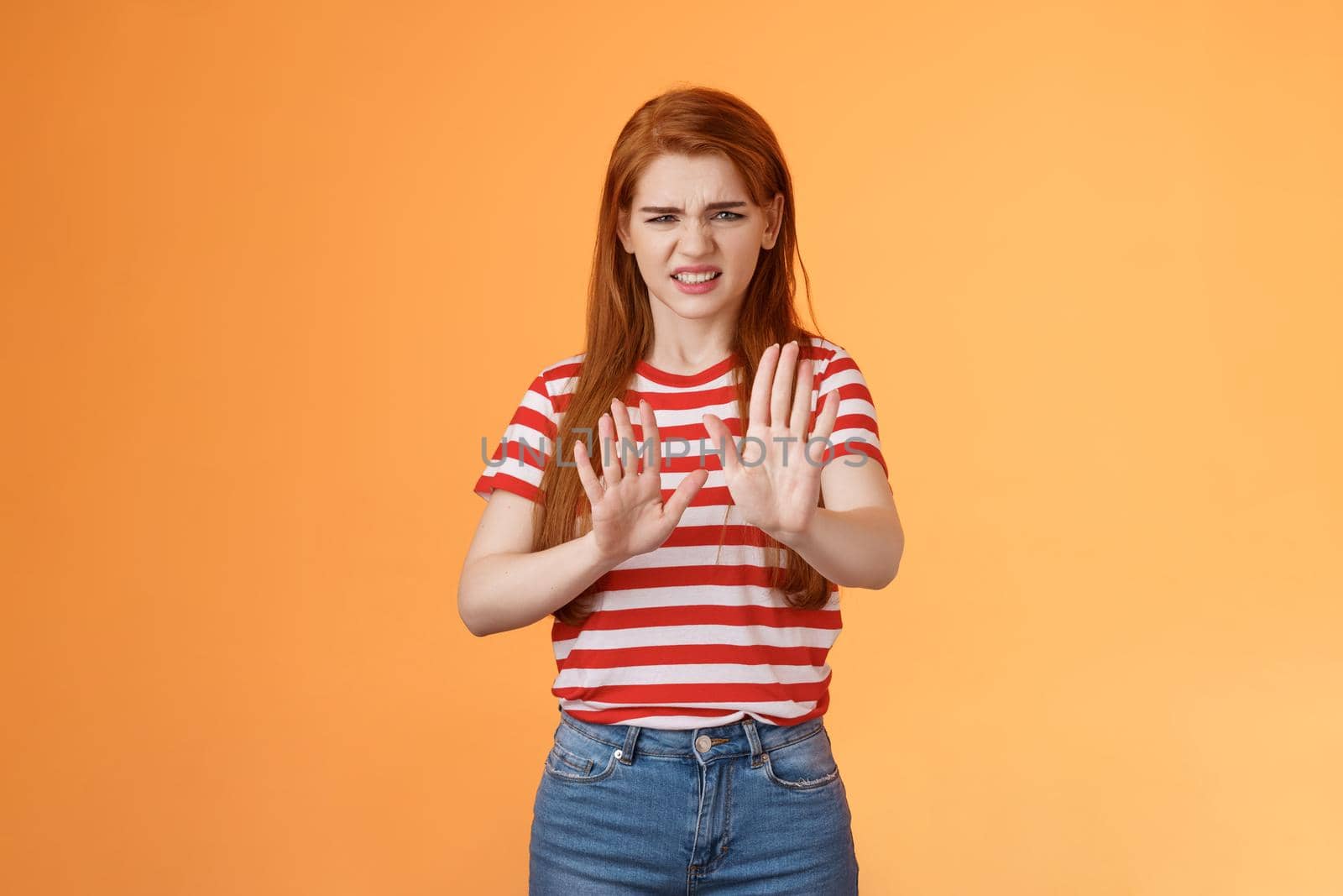 Gosh it stinks. Disgusted redhead picky woman blocking sign raise hands up defensive, grimacing, cringe from aversion awful smell, show refusal rejecting disgusting offer, stand orange background.