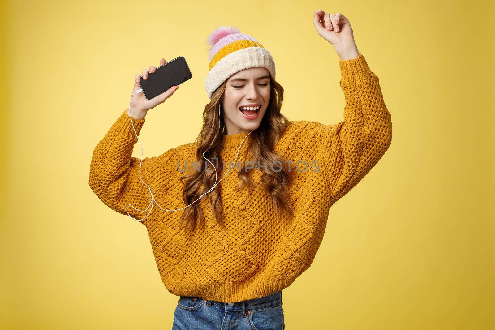 Carefree happy girl enjoying listen music wearing wired earphones raising hands dancing joyfully having fun singing along awesome song playing playlist holding smartphone, standing yellow background by Benzoix