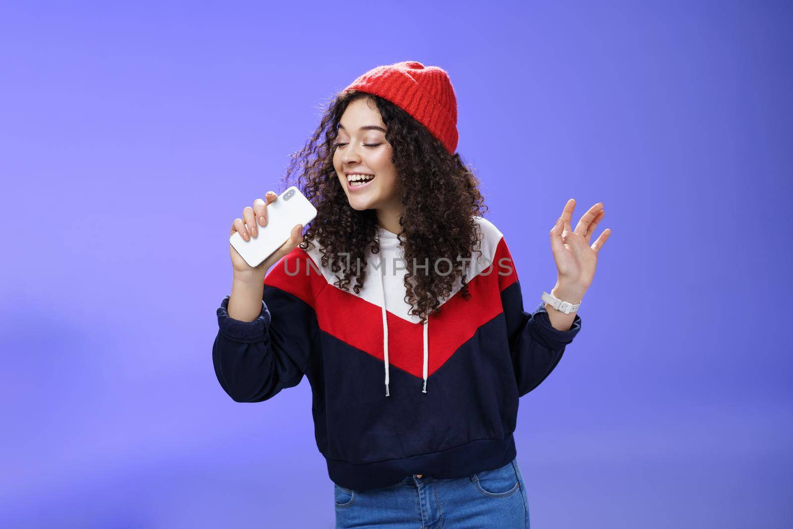 Attractive happy young woman with curly hair in hat singing enjoying perfect winter day singing along in smartphone, holding mobile phone like microphone, adore karaoke over blue background by Benzoix