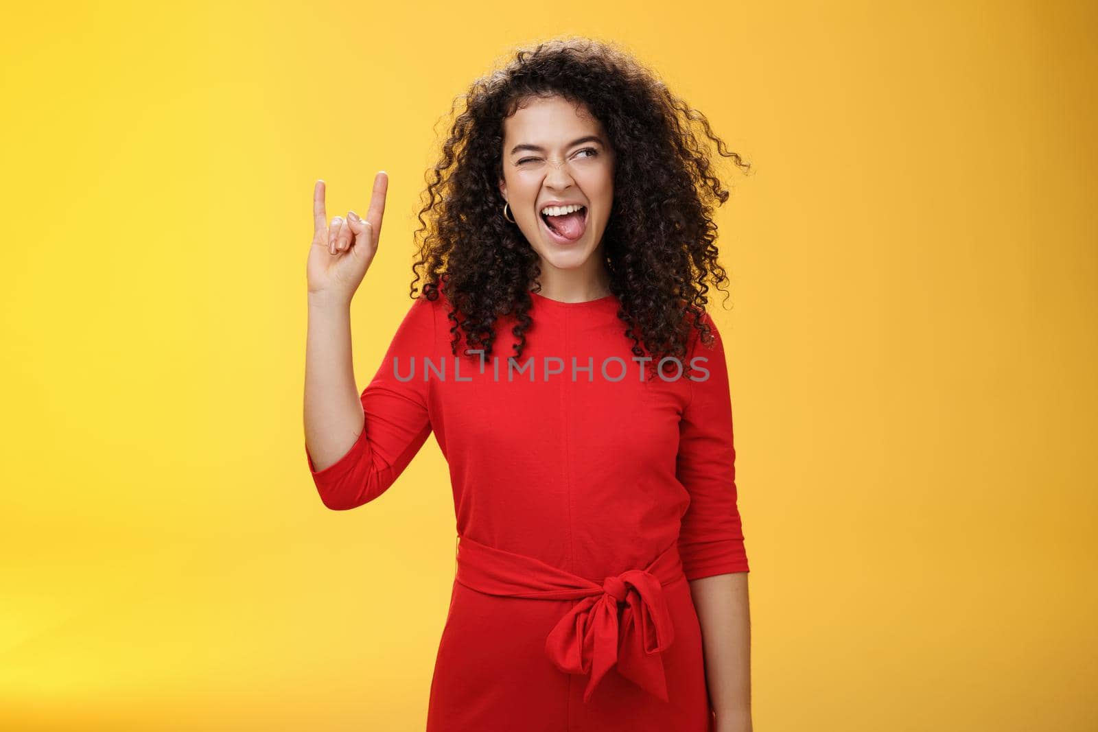 Rebellious meloman leaving in tender girl. Thrilled and carefree curly-haired woman in red dress sticking out tongue and looking right with smile as showing rock-n-roll gesture, enjoying cool music by Benzoix