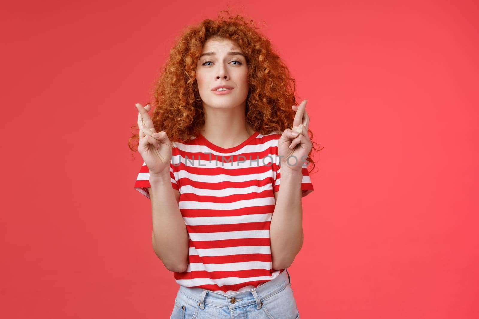 Nervous young silly timid cute redhead ginger girl anticipating hopeful results believe praying squinting intense worry to win cross fingers good luck wish come true red background.