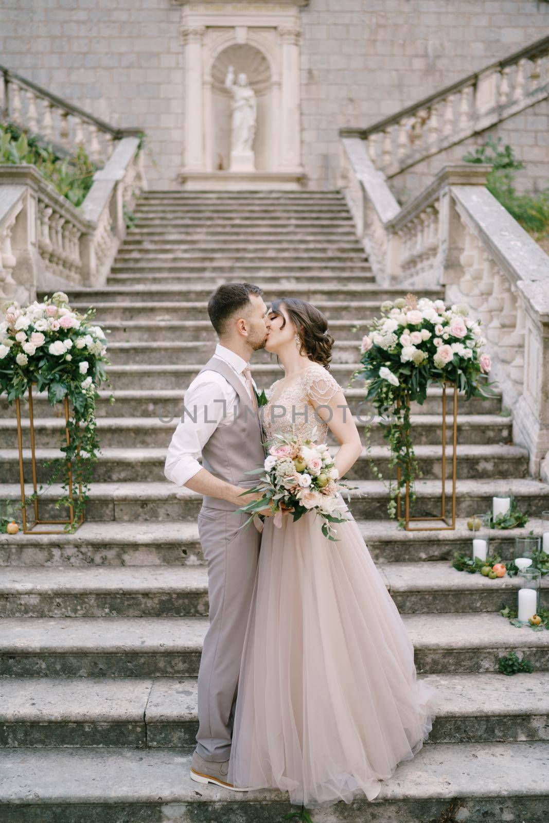 Groom kisses bride while standing on the stone steps near the old villa by Nadtochiy