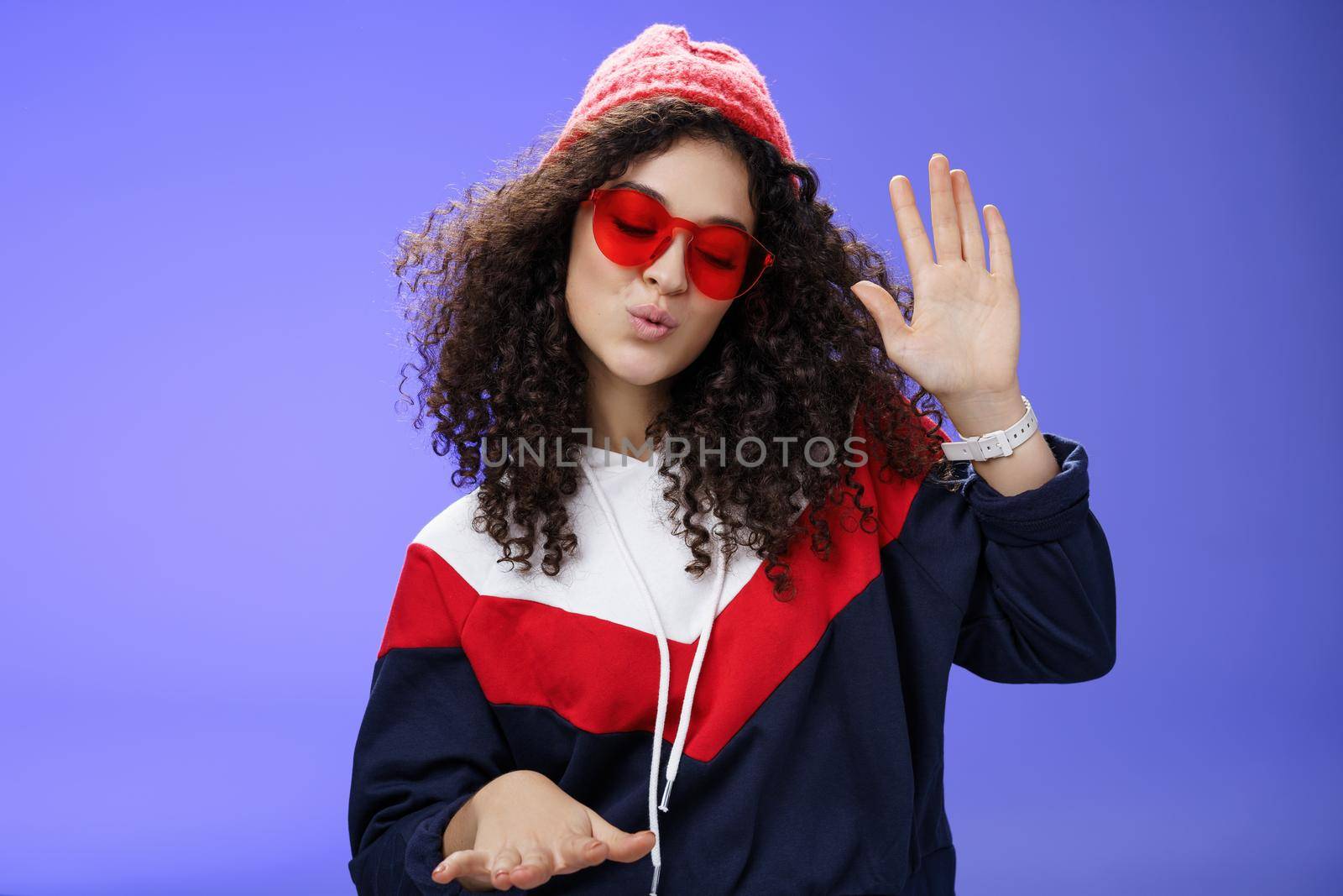 Studio shot of cool and stylish dj girl in red beanie and sunglasses raising palm as enjoying cool music and dancing to rhythm folding lips having fun at awesome party posing chill over blue wall.