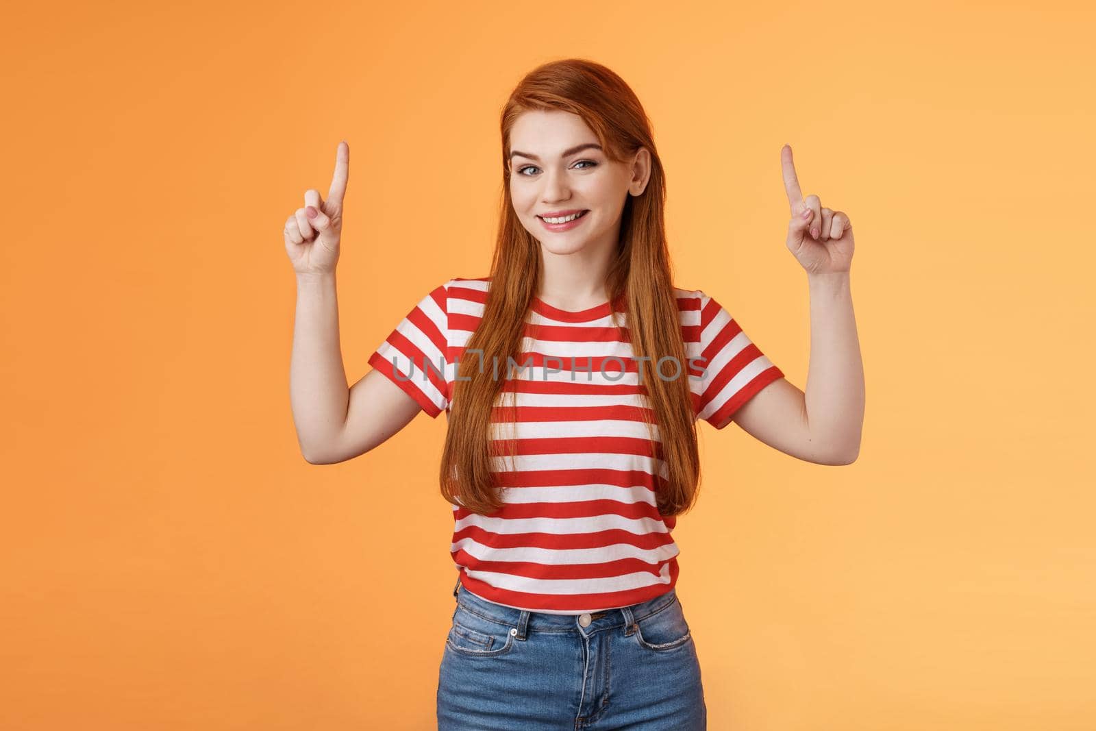 Cheerful beautiful redhead woman smiling invite try out new product, pointing up, show top copy space, grinning joyfully, rejoice introduce cool thing, stand orange background casual urban outfit.