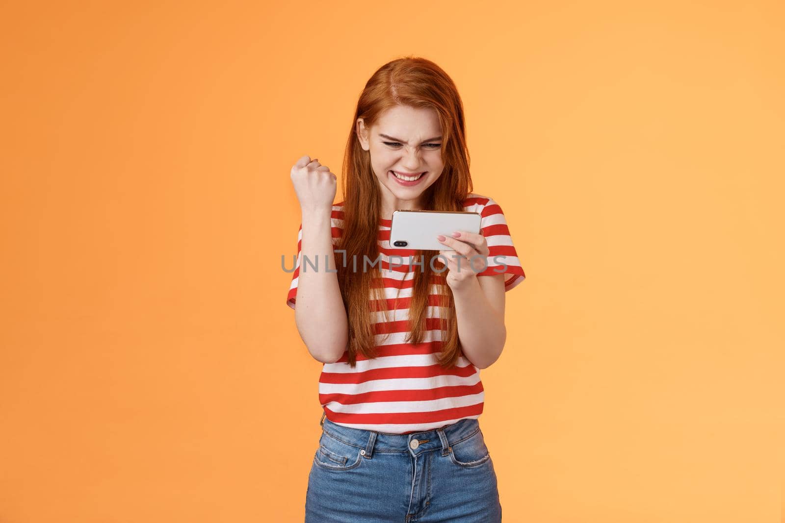 Pleased lucky redhead girl gamer redhead, fist pump satisfied, say yes excited stare display, hold smartphone horizontal, pass hard level playing awesome game use application, achieve victory by Benzoix