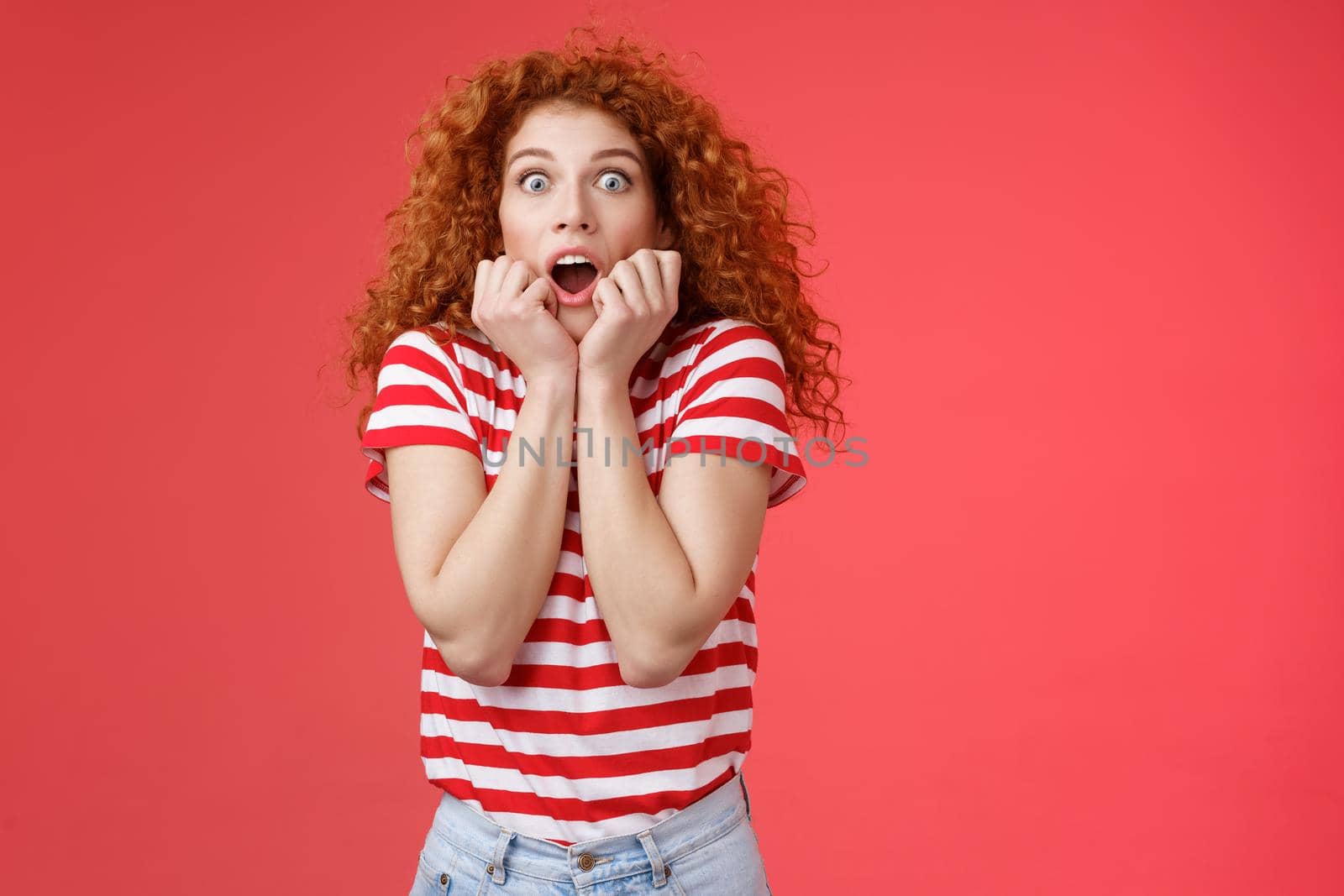 Shocked scared ambushed redhead curly-haired cute girlfriend insecure gasping drop jaw scream frightened stare camera hold hands opened mouth terrified standing stupor red background.