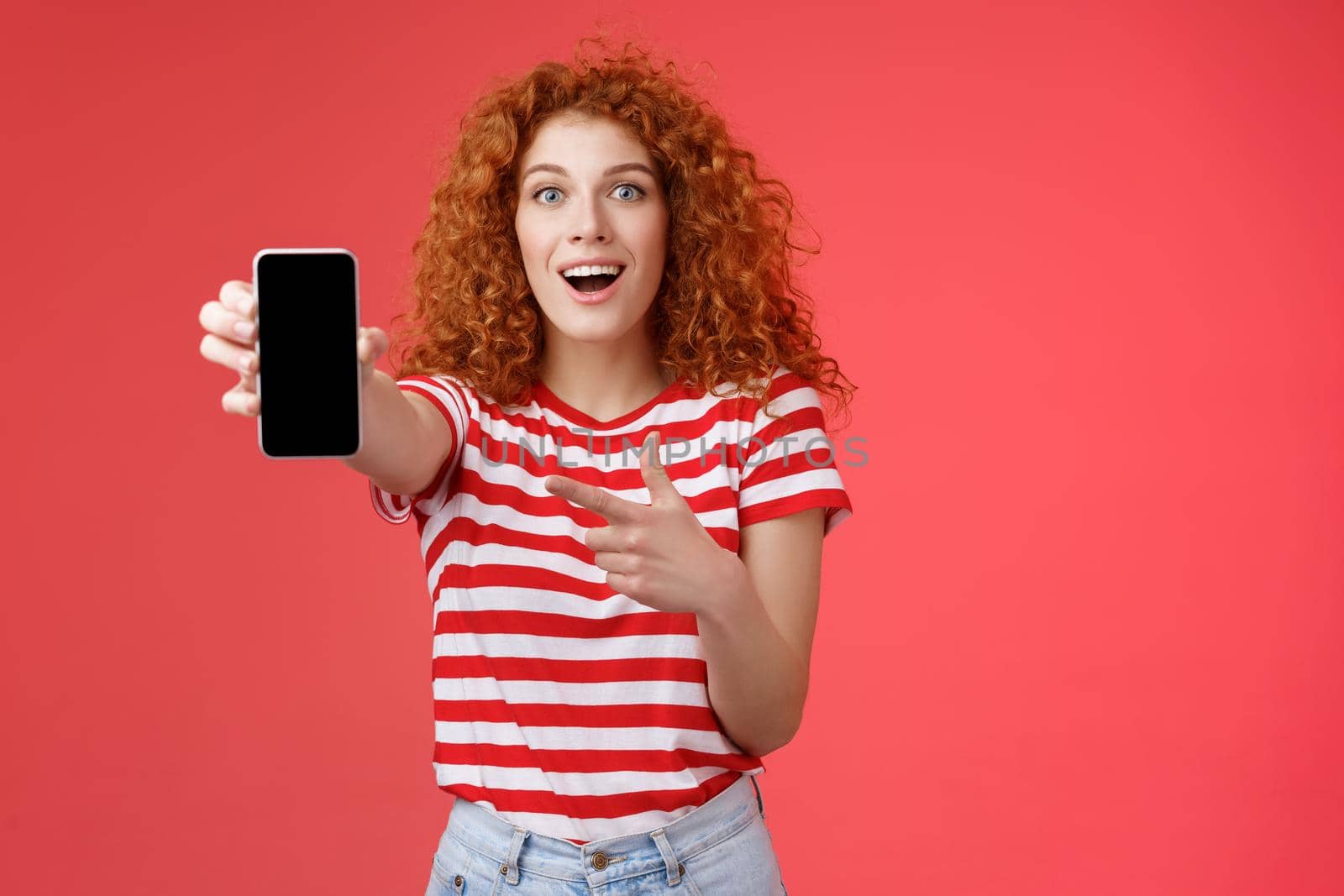 Take look what awesome smartphone. Impressed excited good-looking female love digital innovations redhead girl curly hairstyle showing phone screen pointing display promote cool app.