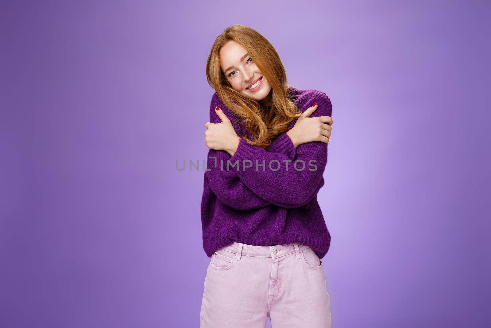 Silly and cute carefree redhead woman in 20s leaning on shoulder as hugging herself feeling warmth wearing purple sweater smiling broadly in cozy and relaxing atmosphere over violet wall by Benzoix
