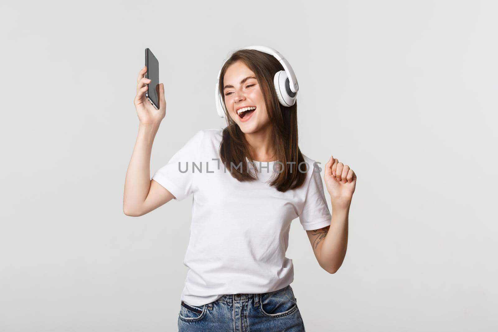 Happy beautiful brunette girl dancing and listening music in wireless headphones, holding smartphone.