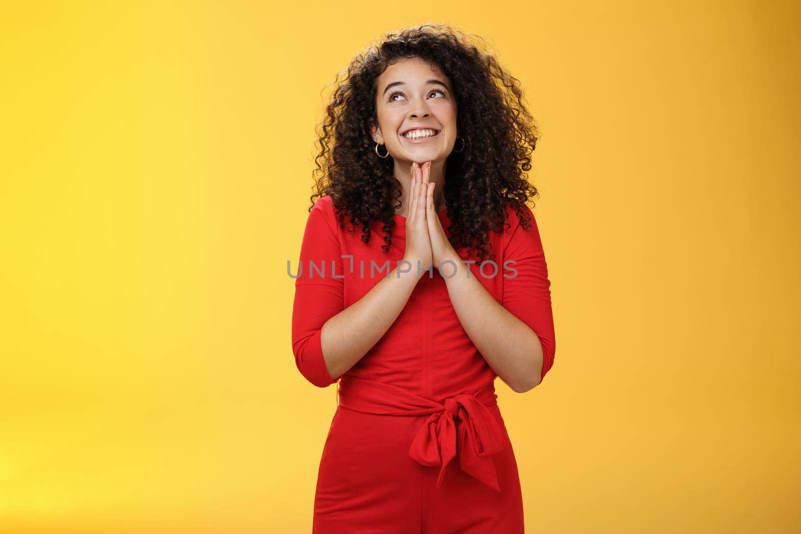 Dreamy excited cute b-day girl with curly hair in cute red dress rubbing palms together near chest as hands in pray smiling looking up delighted and hopeful making wish over yellow background.