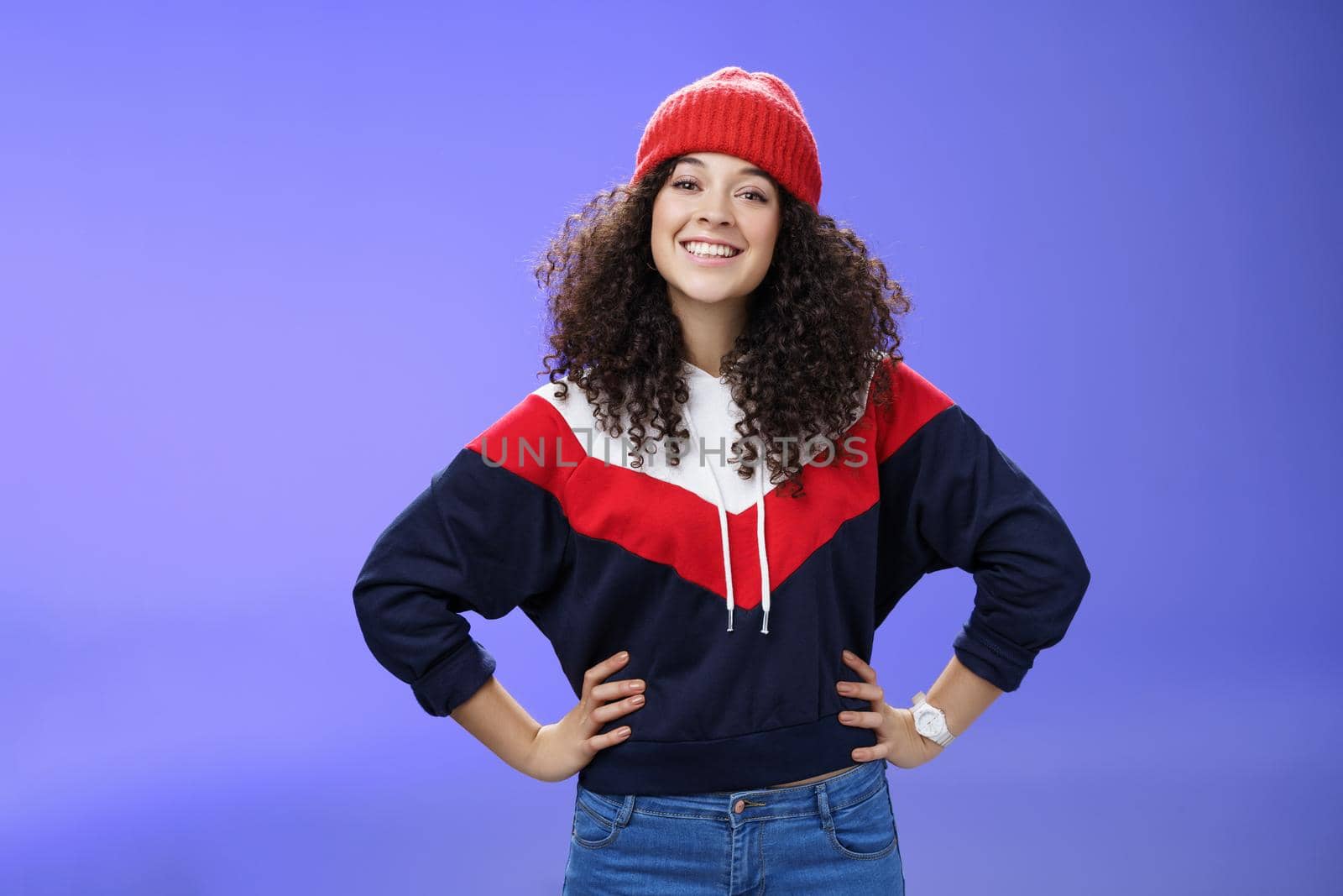 Confident ad energized cute european woman with curly hairstyle in warm beanie and stylish sweatshirt holding hands on waist and smiling friendly at camera as ready to go out and have fun outside by Benzoix