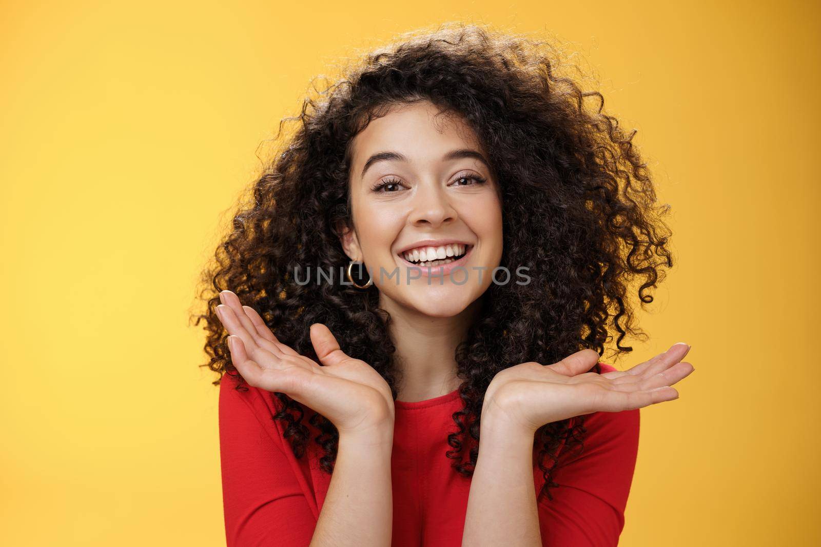 Close-up shot of happy kind and tender pretty caucasian female student with curly hair and perfect skin smiling delighted holding palms spread near face having fun over yellow background.