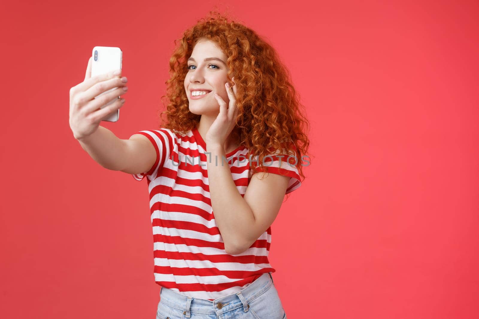 Popular cheerful good-looking stylish female blogger redhead curly hairstyle feel pretty self-acceptance taking selfie raised arm holding smartphone posing silly cute phone camera red background by Benzoix