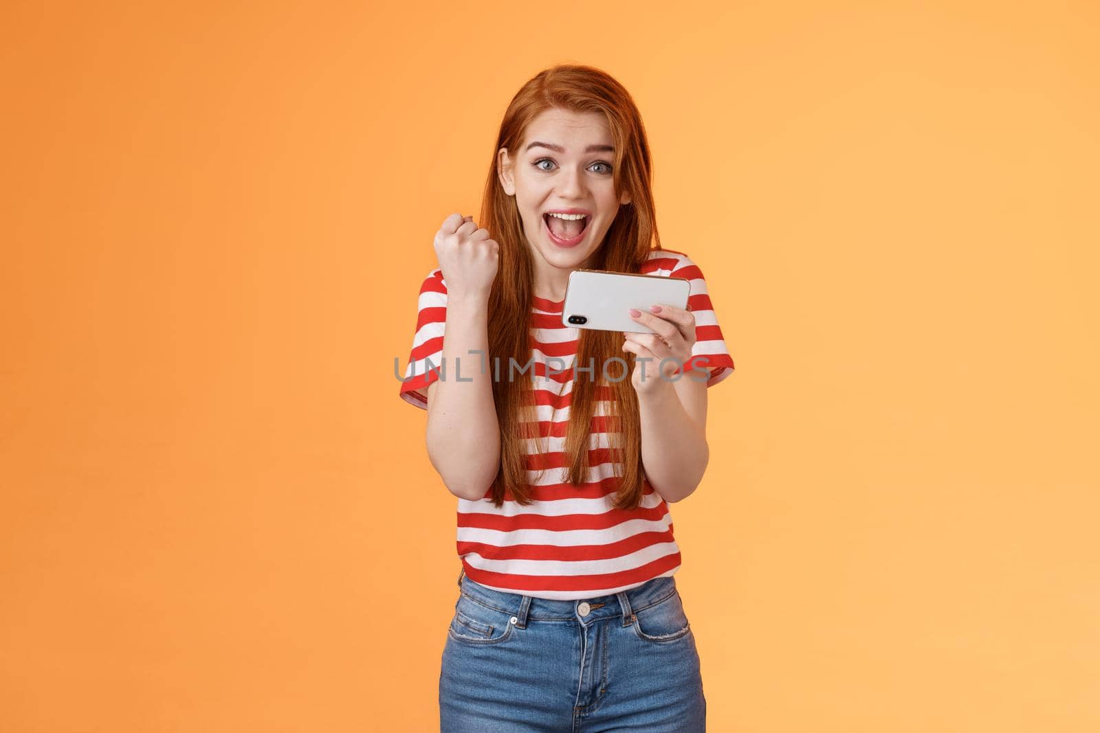 Happy excited redhead female pass level, like awesome game, score goal, hold smartphone horizontally, fist pump success, celebrate lucky achievement, smiling broadly, orange background.