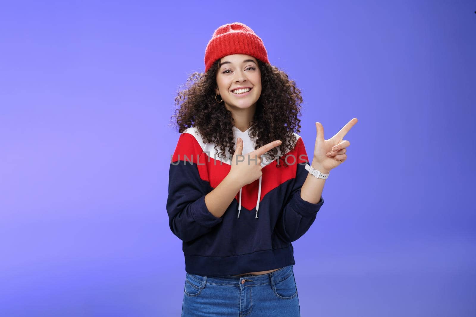 Cute female friend pointing at upper right corner with finger pistols as smiling broadly at camera wearing warm winter hat and sweatshirt skiing over blue background joyfully by Benzoix