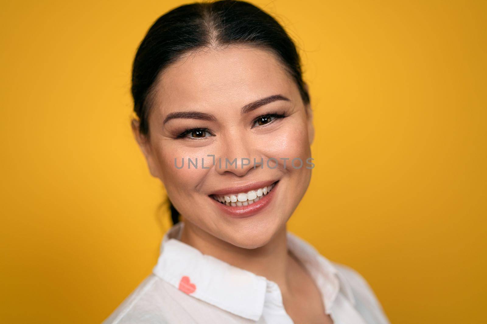 Pleasant looking glad Asian woman looks in camera, has toothy smile, wears casual clothing, isolated on yellow background. Human emotions, facial expression concept. Facial expressions, feelings.