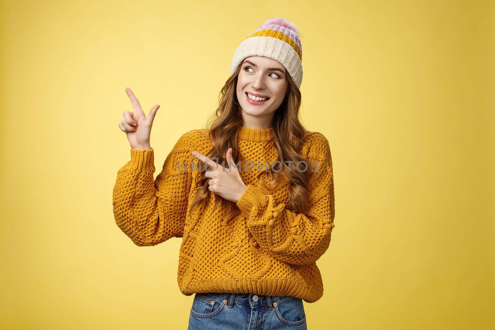 Curious attractive young 20s woman intrigued what hiding left pointing sideways looking interested smiling excited peeking cool fantastic promotion, standing joyfully yellow background. Copy space