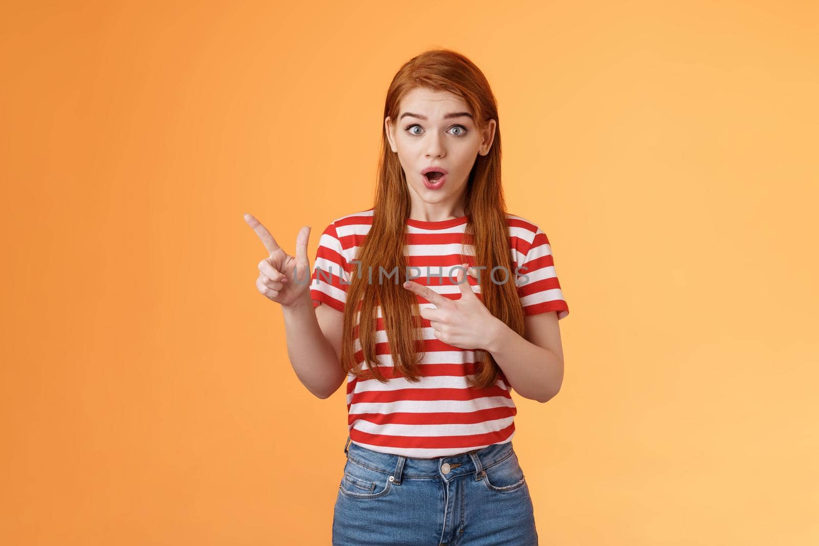 Wow awesome. Impressed excited beautiful redhead woman look astonished explain amazing news, stare camera thrilled talking about advertisement, pointing left upbeat impressed, orange background.