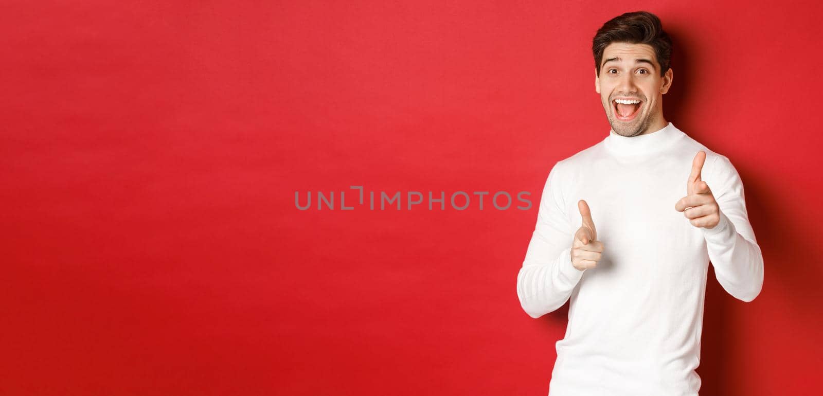 Concept of winter holidays, christmas and lifestyle. Cheerful handsome guy in white sweater congratulating you, pointing fingers at camera and wishing happy new year, standing over red background.