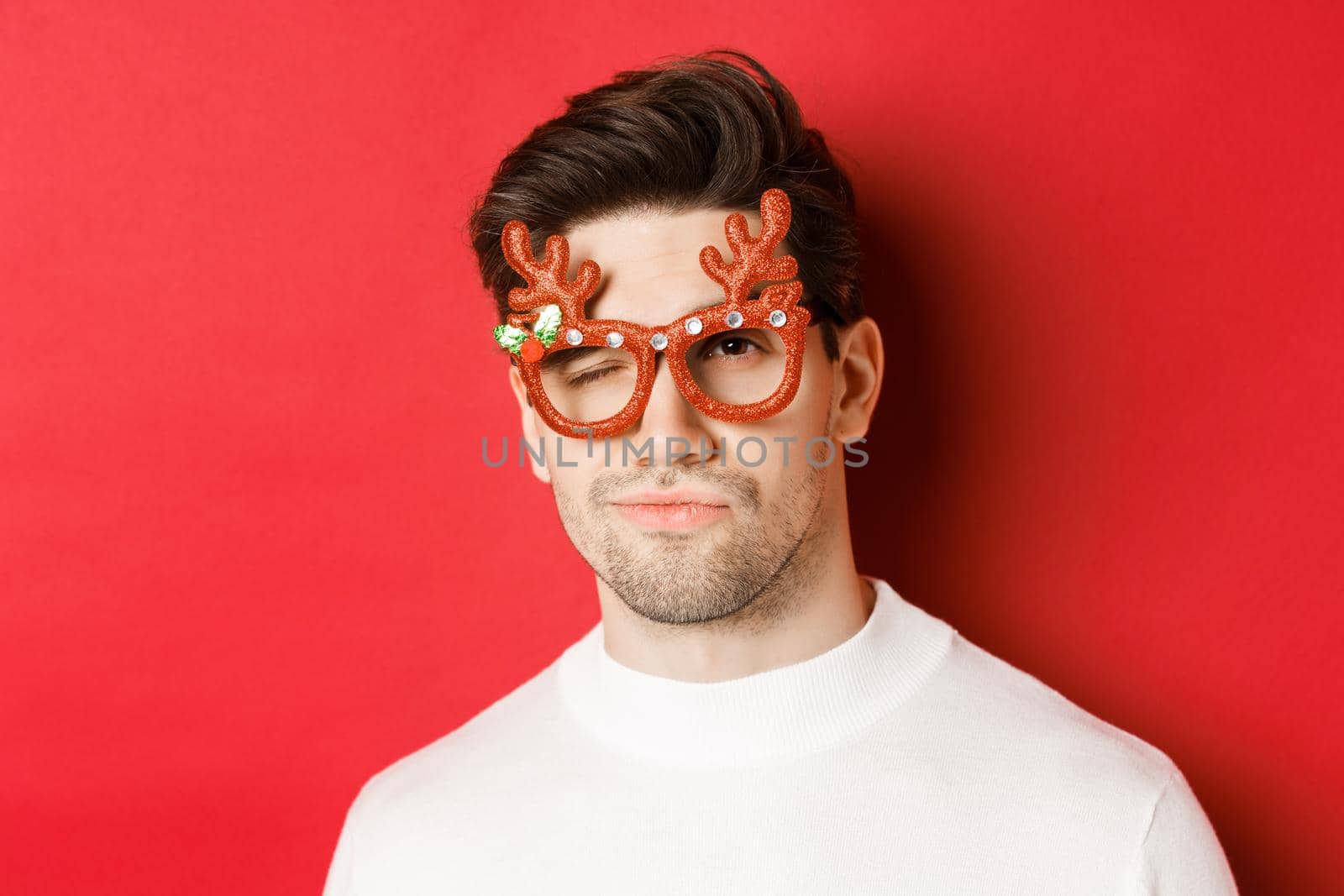 Concept of winter holidays, christmas and celebration. Close-up of cheeky attractive man in party glasses, winking and looking sassy, standing over red background.