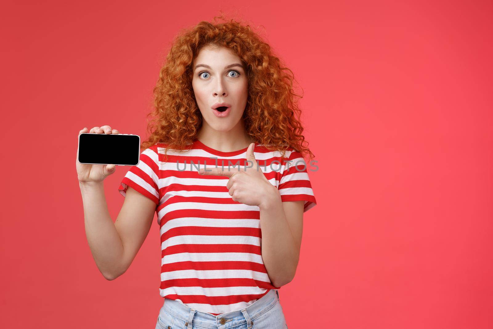 Amused fascinated cute redhead curly girl adore playing games hold smartphone horizontal show thumb up open mouth gasping amazed happy cheerfully present own score favorite app red background by Benzoix