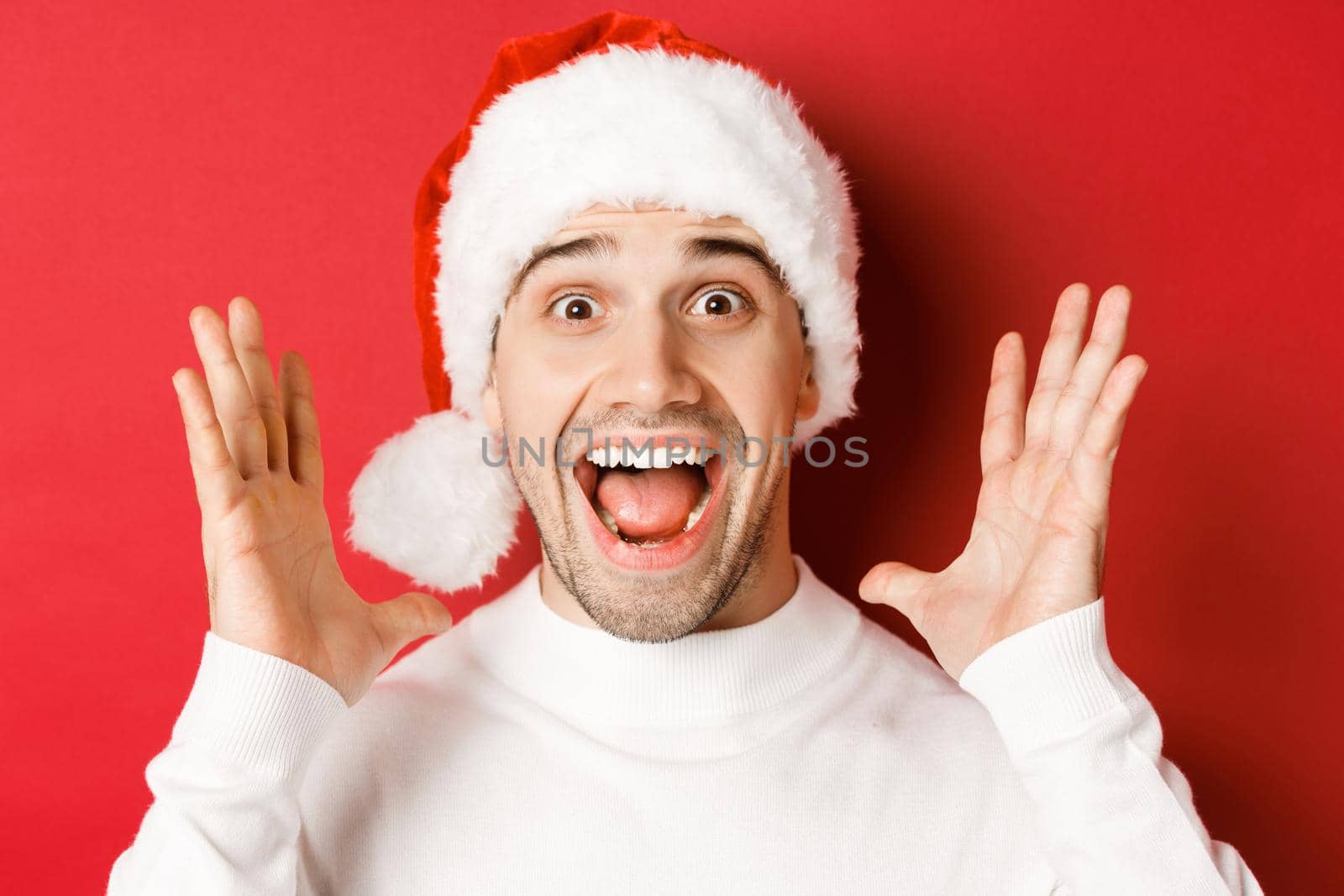 Close-up of happy young man in santa hat, making big christmas announcement, smiling amazed, standing over red background.
