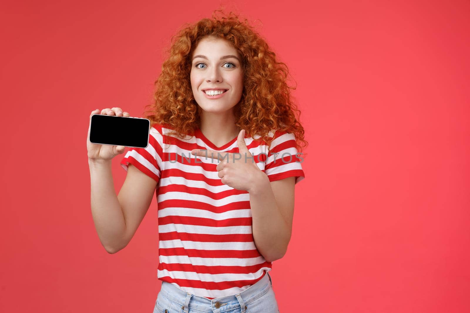Excited happy good-looking european redhead curly girl amazed showing personal achievement score smartphone game show phone screen pointing display discuss awesome gameplay smiling delighted.