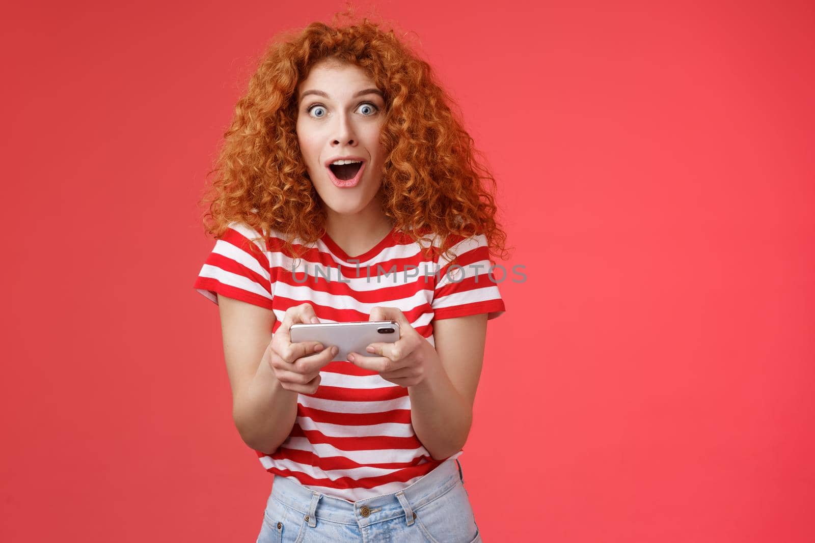 Impressed excited good-looking redhead curly woman hold smartphone horizontal open mouth fascinated stare camera thrilled astonished win passed level game happy playing cool app red background.