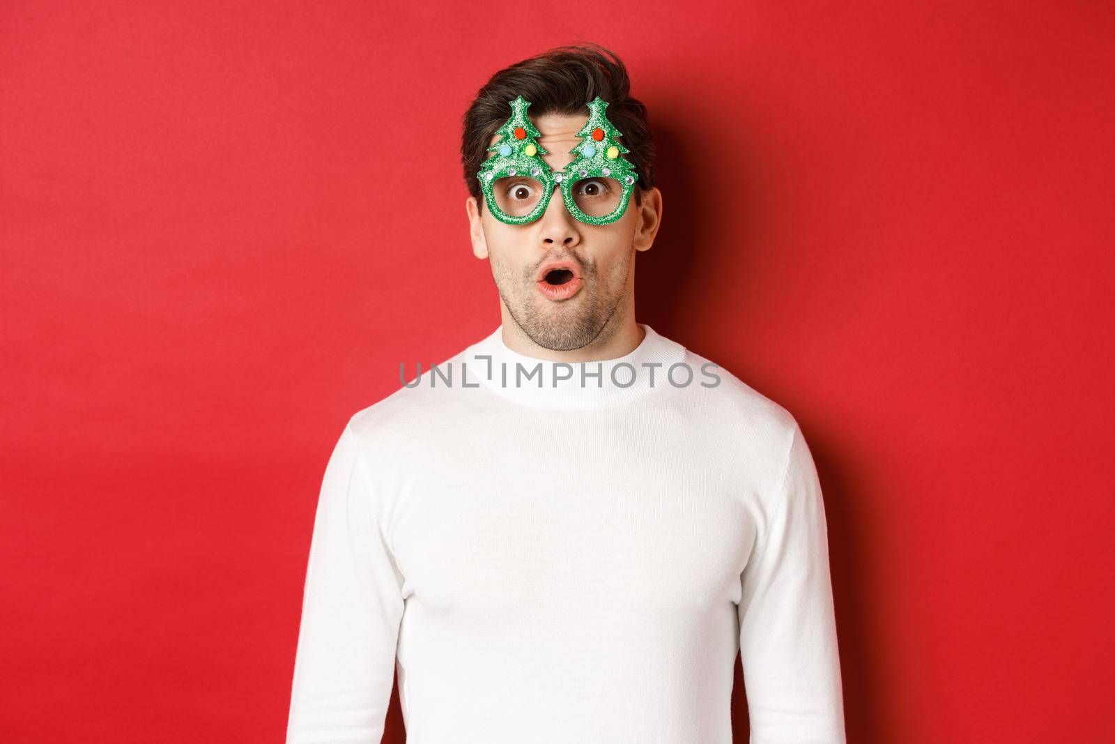 Concept of christmas, winter holidays and celebration. Close-up of surprised handsome guy in party glasses, saying wow and looking at amazing new year promo offer, red background.