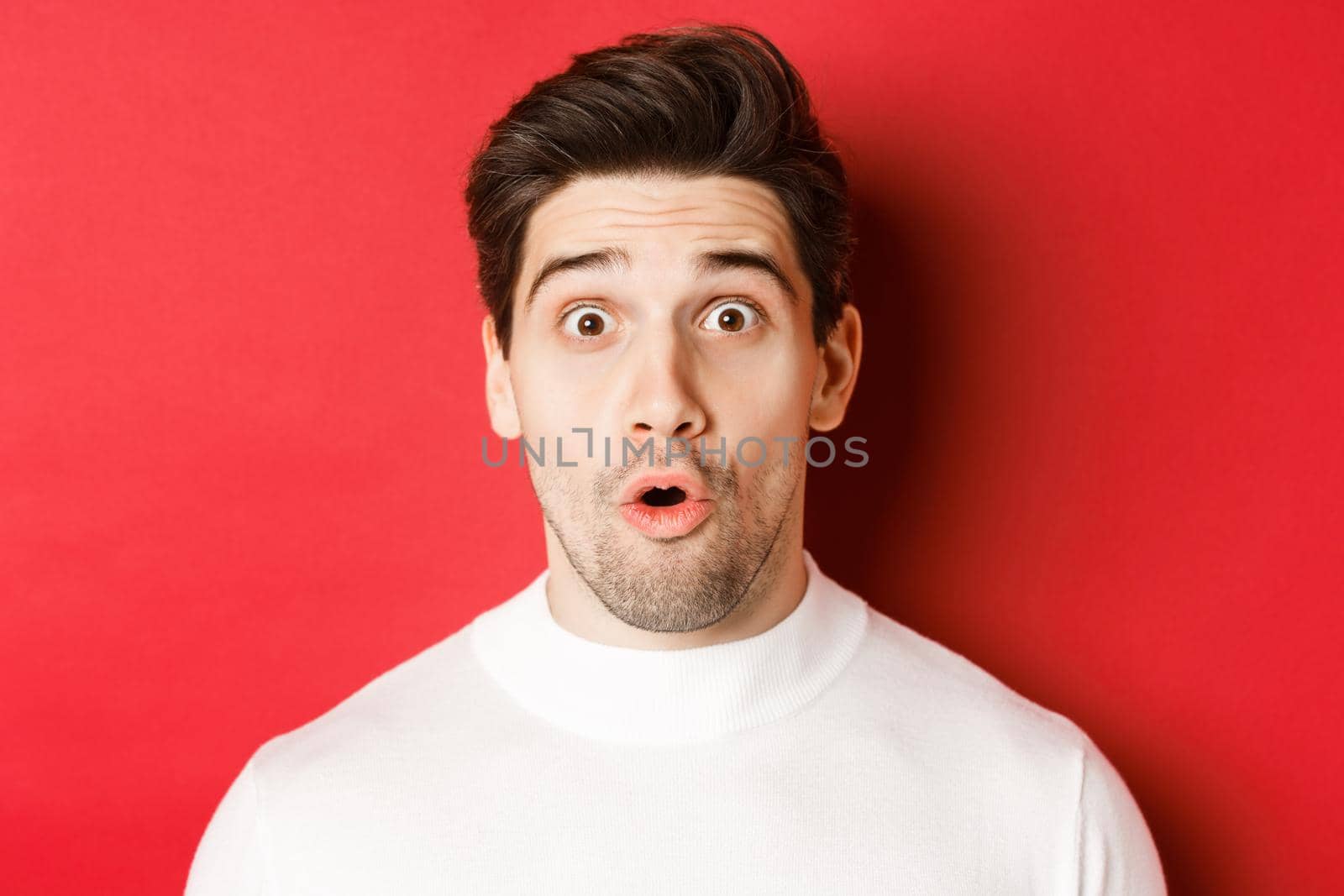 Close-up of surprised handsome man in white sweater, open mouth and looking wondered, hear amazing news, standing over red background.