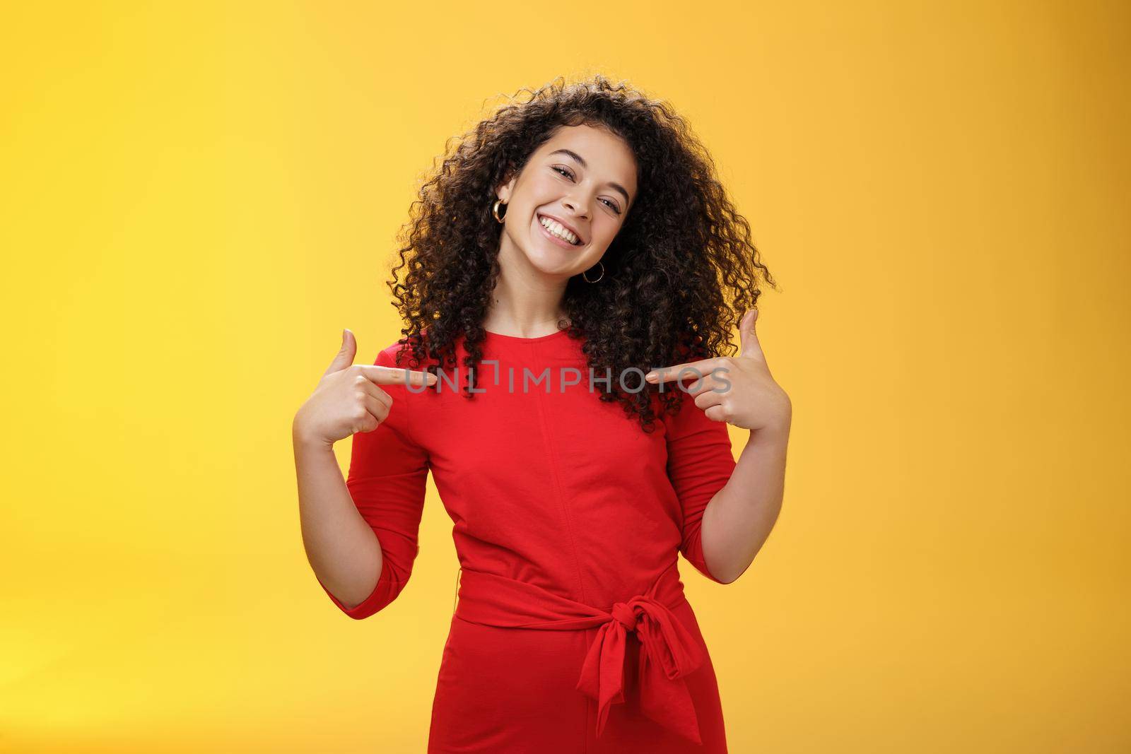 Proud and satisfied ambitious successful female student in red dress standing pleased smiling and pointing at herself as if bragging about own achievements happily and glad over yellow wall.