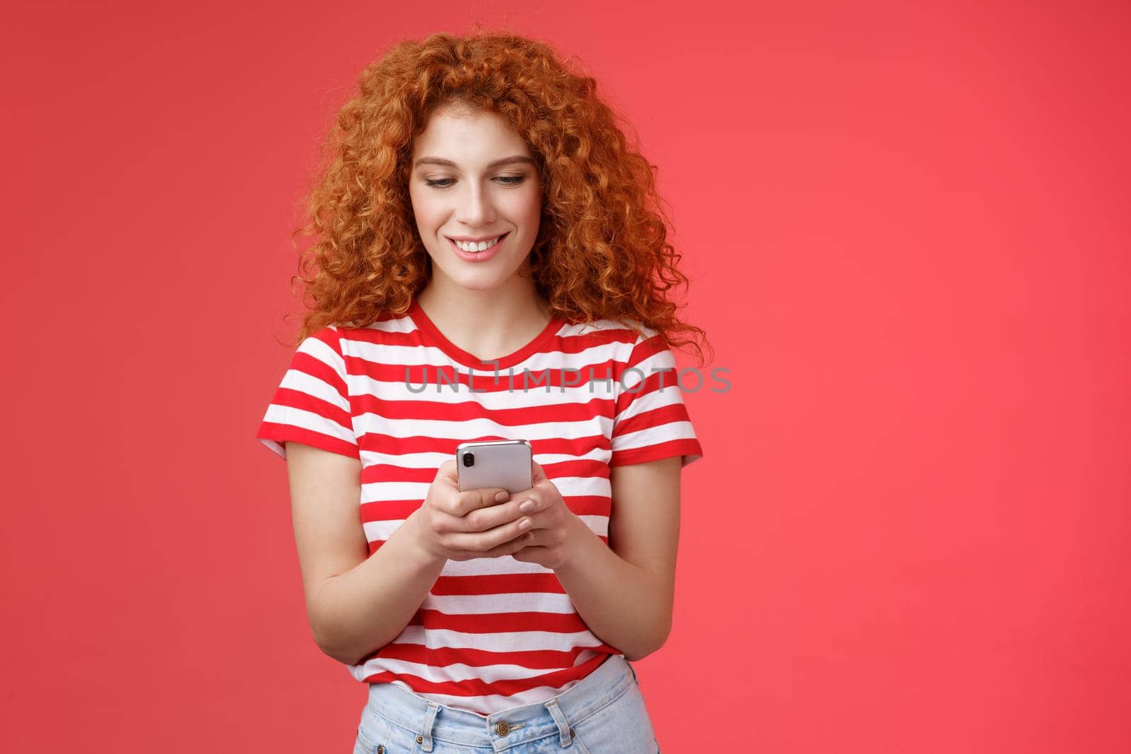 Time check social media feed. Good-looking relaxed happy fashionable redhead woman curly hairstyle hold smartphone look phone screen amused smiling delighted texting friend throw party asap by Benzoix