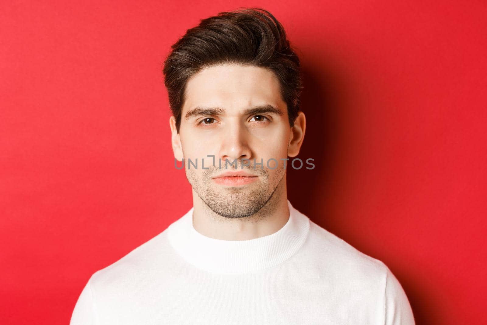 Close-up of thoughtful, serious-looking man in white sweater, standing over red background.