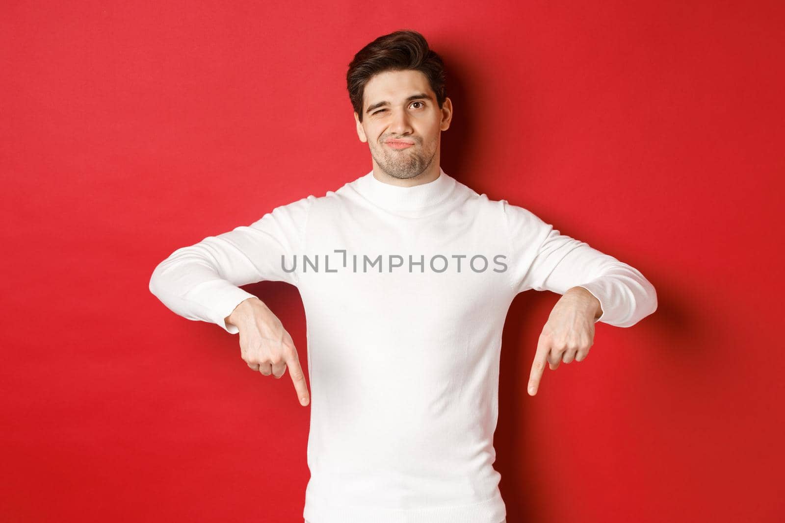 Portrait of attractive, skeptical young man in white sweater, grimacing and pointing fingers down, disagree and dislike something, standing over red background.