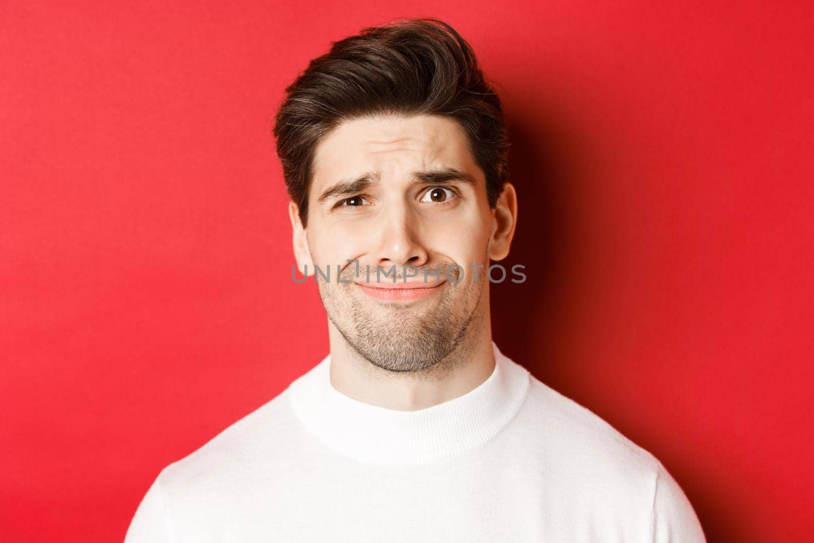 Close-up of handsome man feeling uncomfortable, grimacing and looking at something displeasing, standing over red background.