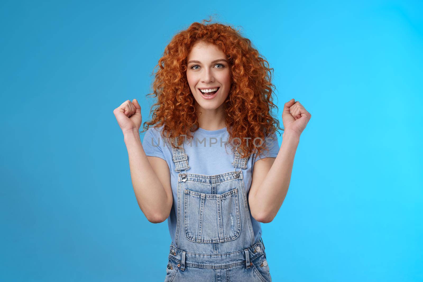 Hooray successful day. Cheerful chrismastic redhead girl summer dungarees clench fists joyfully smiling broadly determined achieve success, triumphing winning game standing upbeat blue background.