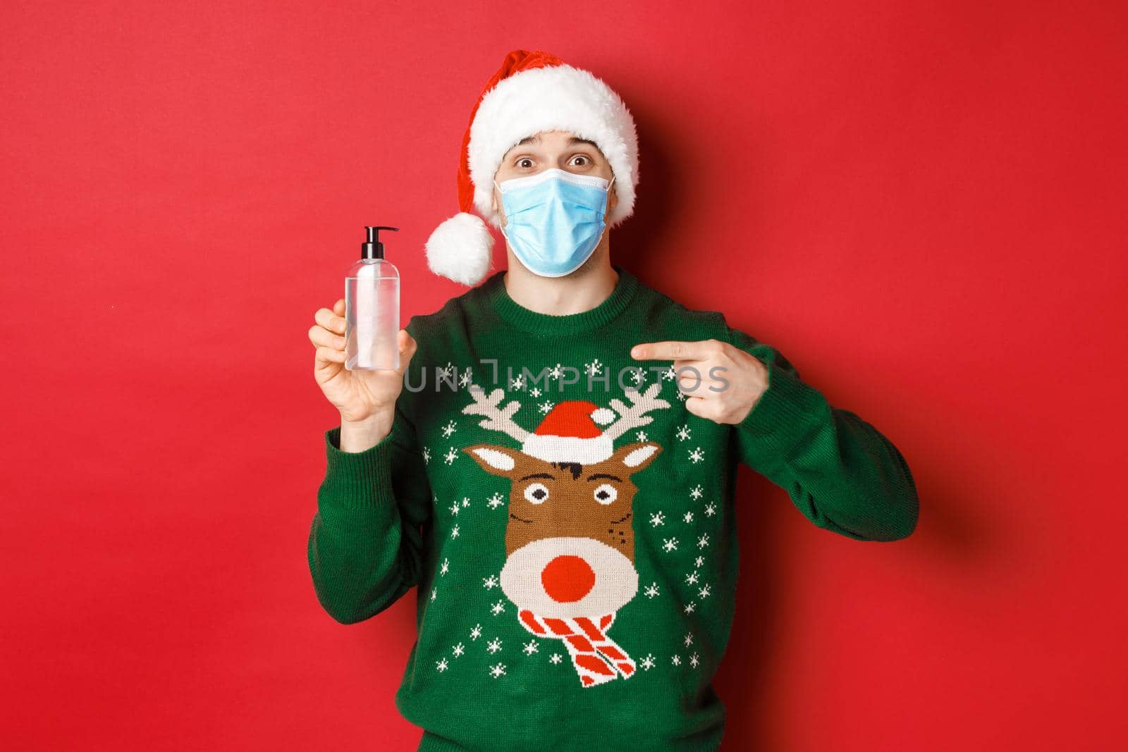 Concept of new year, coronavirus and social distancing. Portrait of cheerful man in santa hat, christmas sweater and medical mask, pointing finger at hand sanitizer, standing over red background.